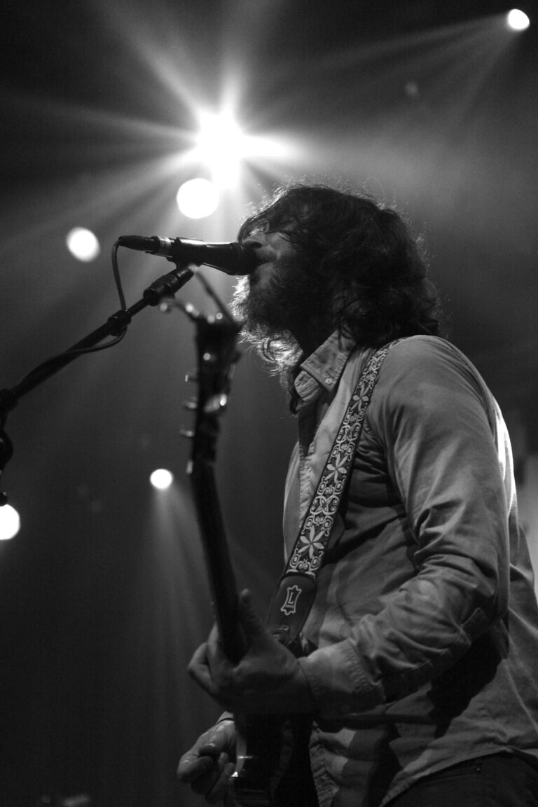 Een muzikant staat op het podium en zingt in een microfoon, met een gitaar in de hand en opvallende lichteffecten op de achtergrond.
