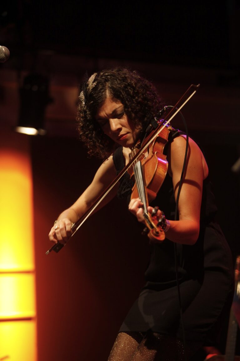 Een vrouwelijke violiste speelt met intensiteit op een podium, omringd door sfeervolle verlichting.