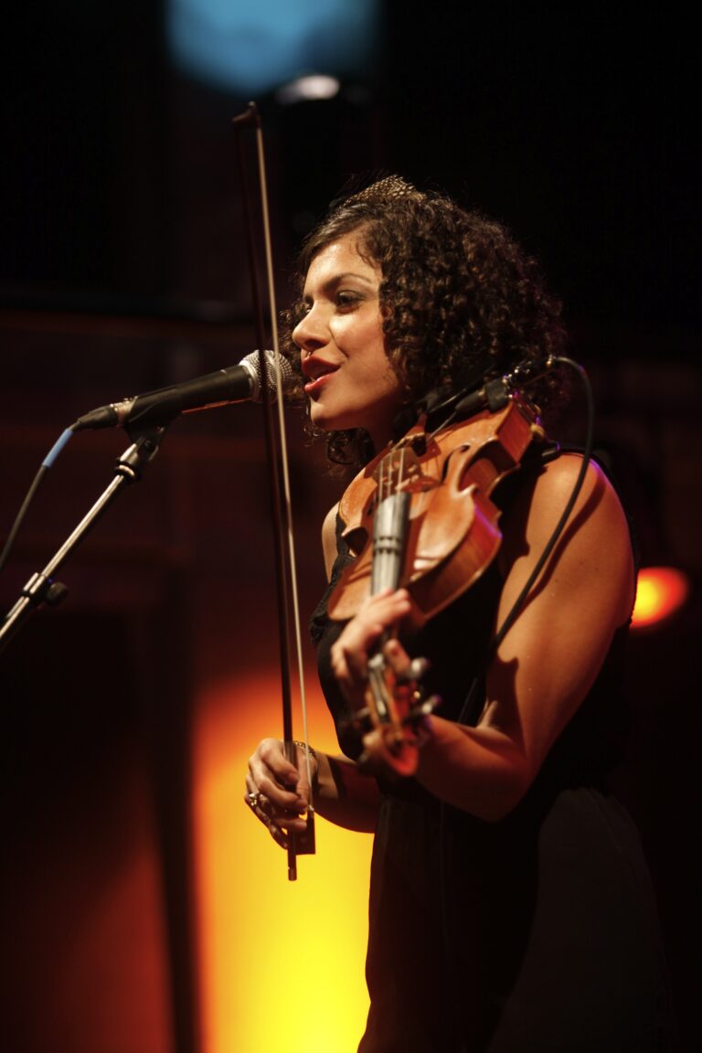 Een muzikante die een viool bespeelt terwijl ze zingt in een sfeervolle podiumomgeving.