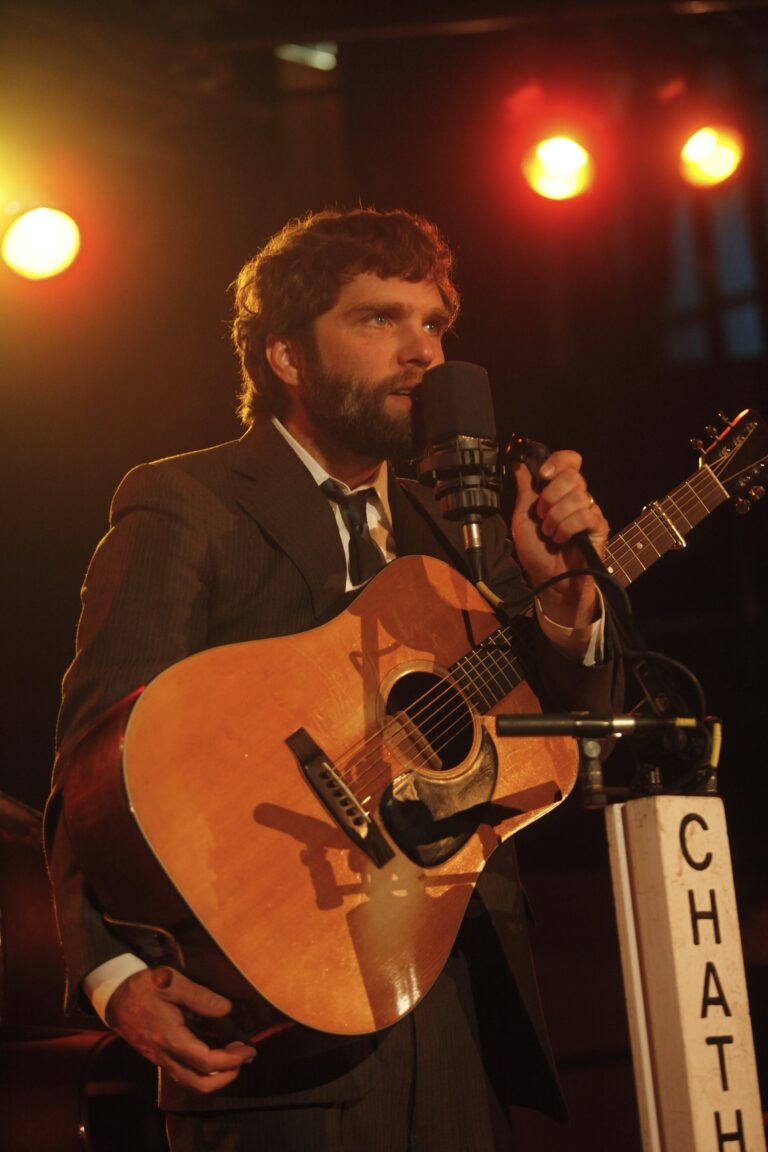 Een man met een gitaar staat achter een microfoon en zingt op een podium met sfeervolle verlichting.