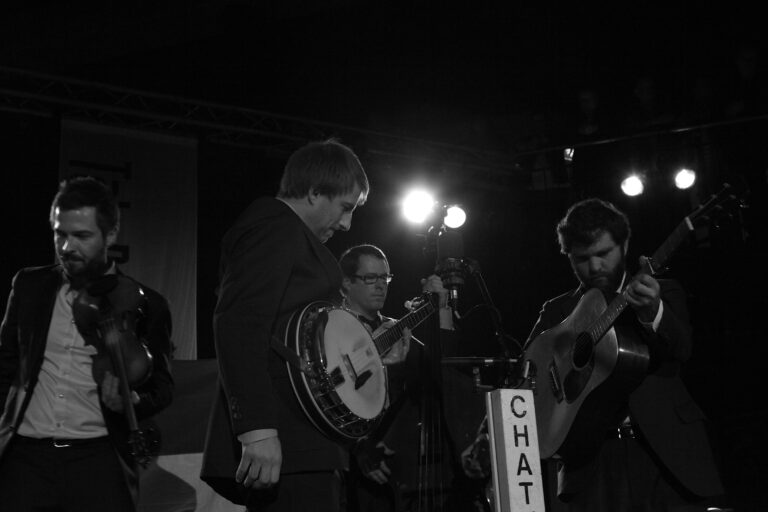 Zwart-witfoto van een live muziekoptreden met muzikanten die verschillende instrumenten bespelen, waaronder een banjo en een gitaar.