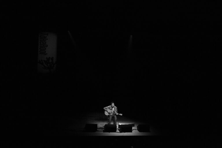 Een muzikant speelt gitaar op een donkere podium, verlicht door enkele lichten.