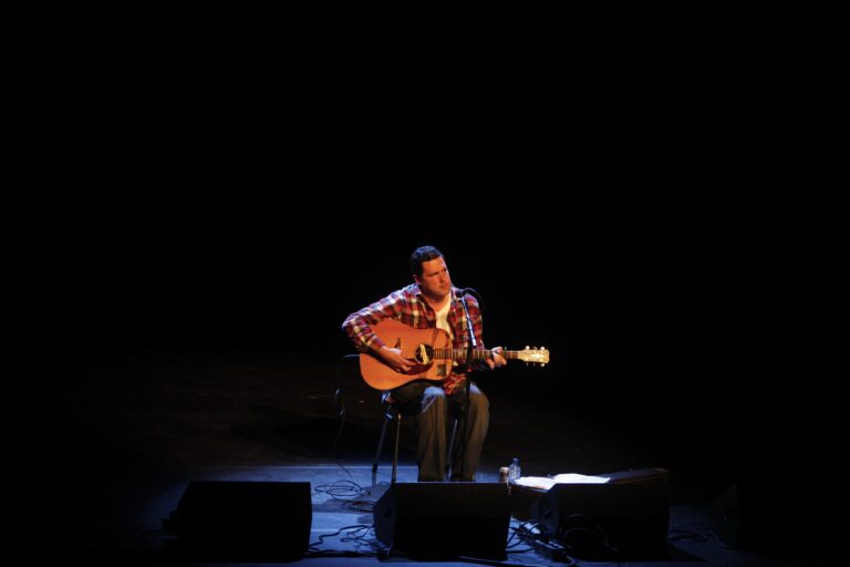 Een muzikant zit op een stoel en speelt gitaar in een donkere omgeving met een enkel licht.