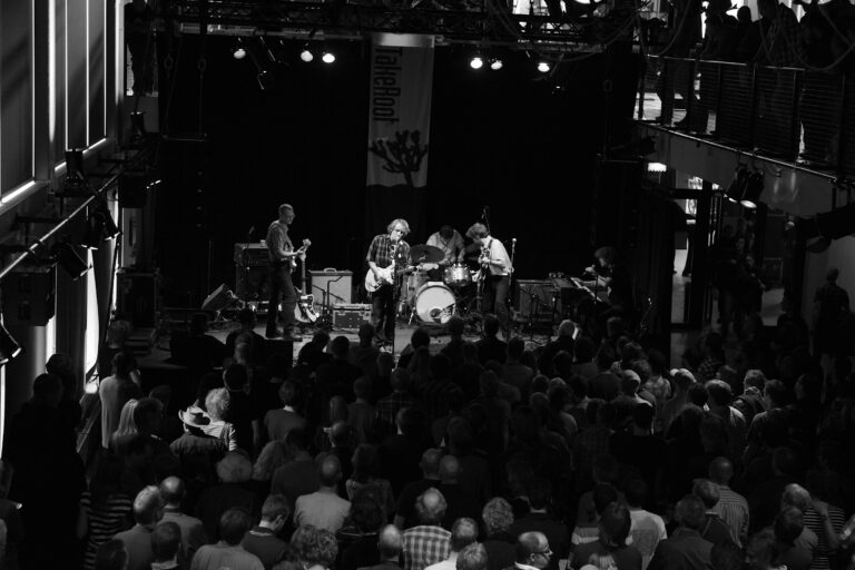 Een zwart-witfoto van een band die live optreedt voor een publiek in een drukke zaal.