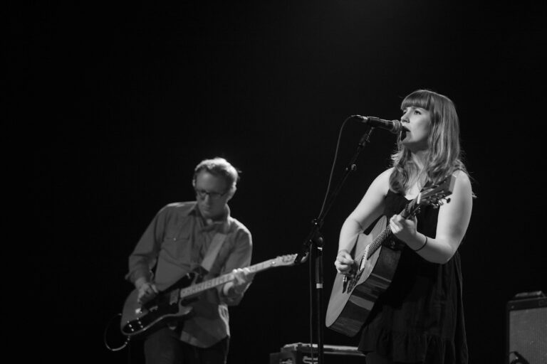 Een zangeres met een gitaar optredend op een podium, terwijl een muzikant op de achtergrond een elektrische gitaar bespeelt.