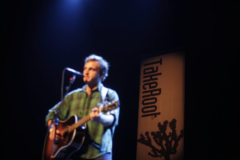 Een muzikant met een gitaar staat op een podium met een lichteffect en een banner op de achtergrond.