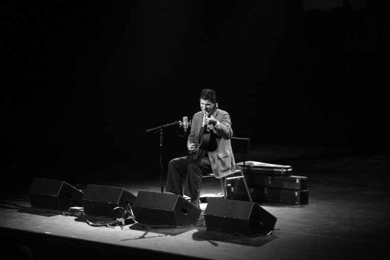 Een muzikant speelt gitaar op een podium, omgeven door microfoons en een donkere achtergrond.