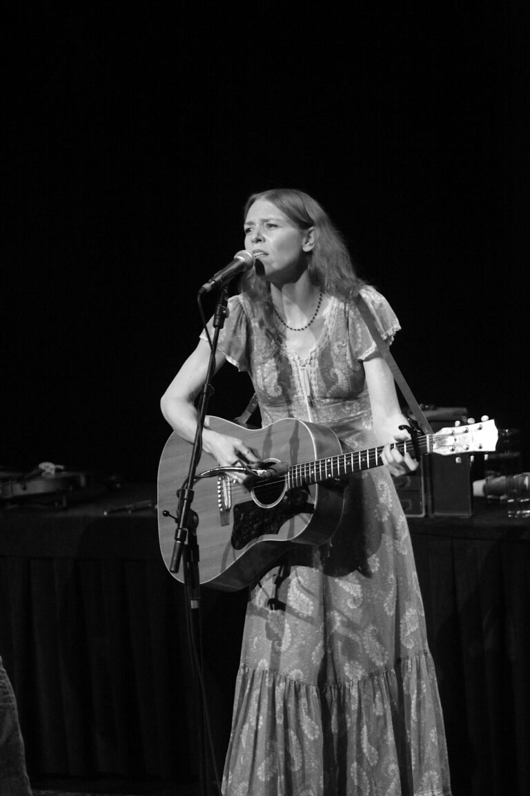 Een vrouw speelt gitaar en zingt op een podium in zwart-wit.