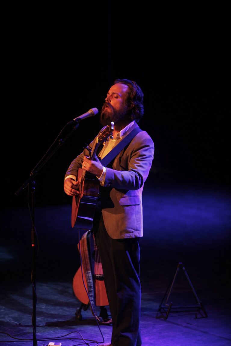 Een man met een baard speelt gitaar en zingt in een sfeervolle, gezellige setting met blauwe verlichting.