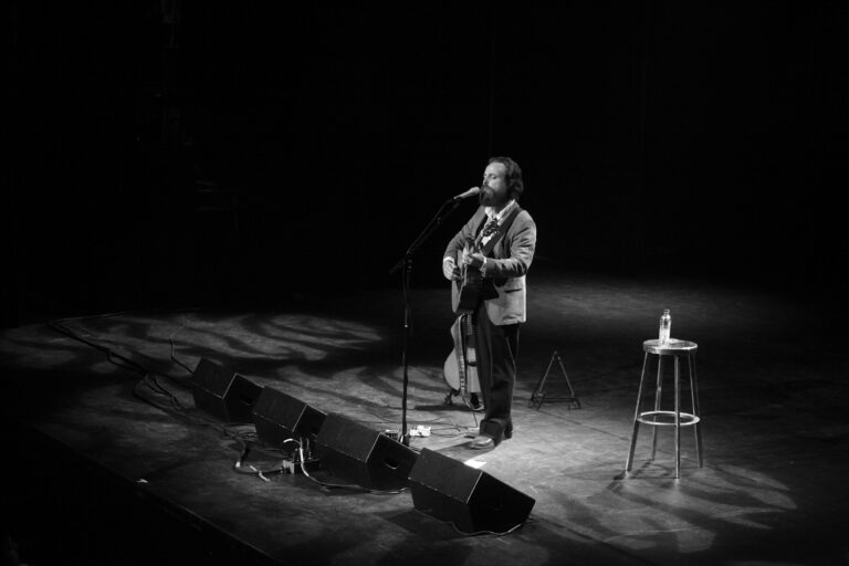 Een muzikant staat op het podium en speelt gitaar, met een microfoon en een kruk naast zich.