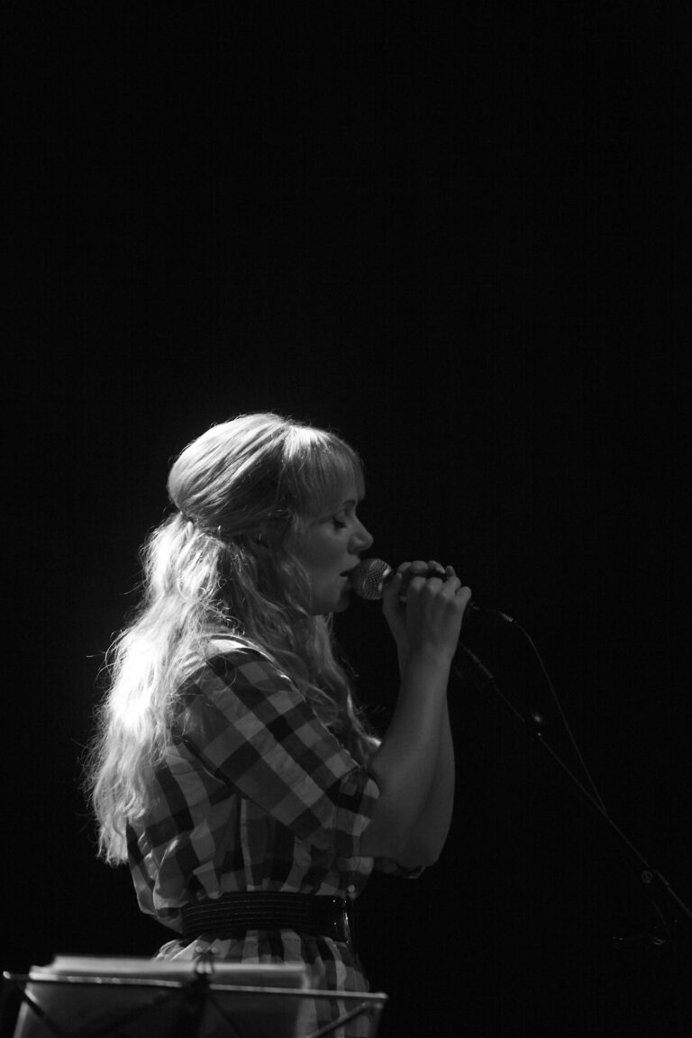 Zijprofiel van een vrouw met lang haar, die in een microfoon spreekt op een donkere achtergrond.