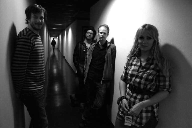 Een z/w afbeelding van vier mensen die in een gang staan, met een silhouet van een vijfde persoon in de achtergrond.