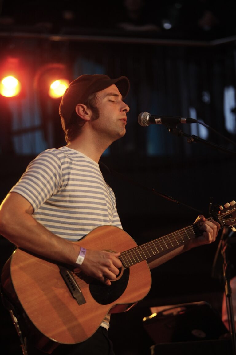 Een man met een gitaar zingt op een podium, verlicht door warme spotlights.