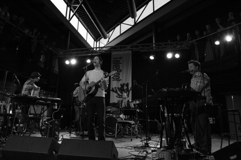 Een band speelt live muziek op een podium met diverse instrumenten en verlichting in een zwarte en witte sfeer.