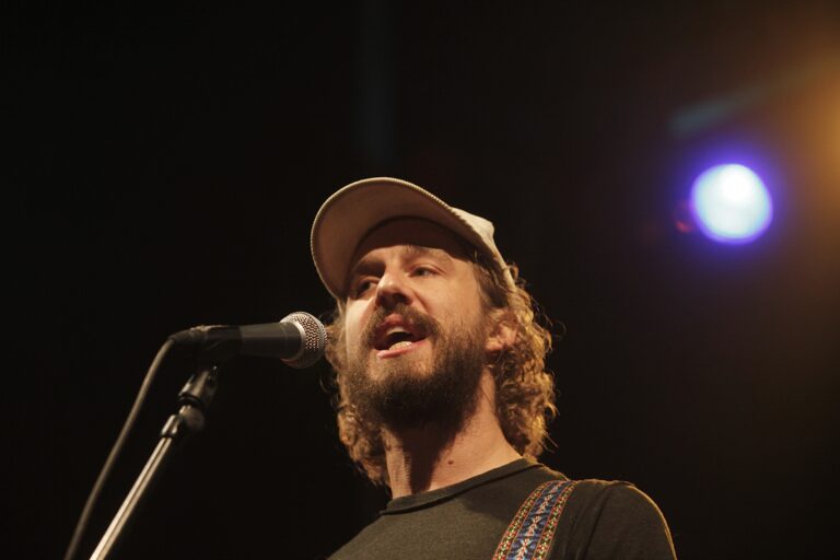 Een zanger met een pet, die in een microfoon zingt, met een podiumverlichting op de achtergrond.