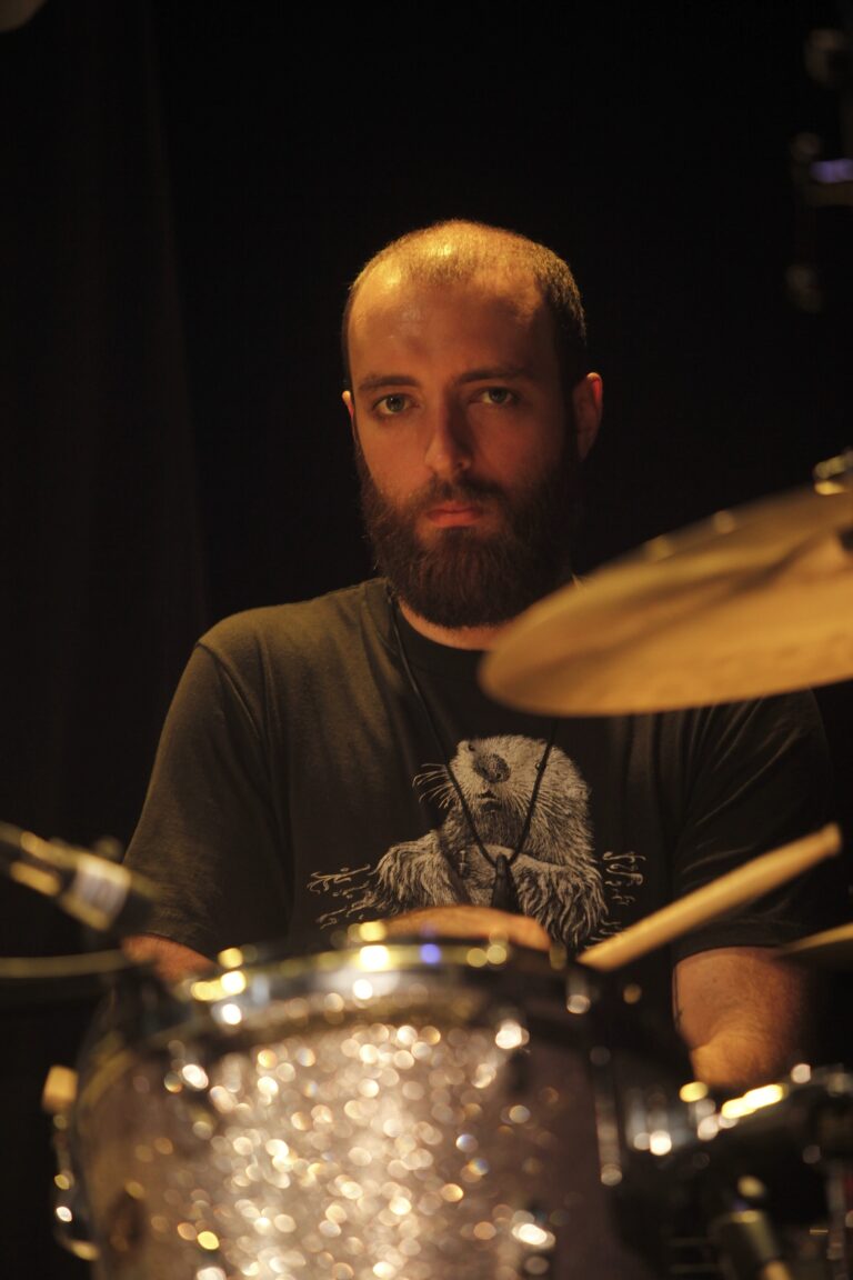 Een drummer speelt op een podium met een geconcentreerde uitdrukking, terwijl hij naar de camera kijkt.