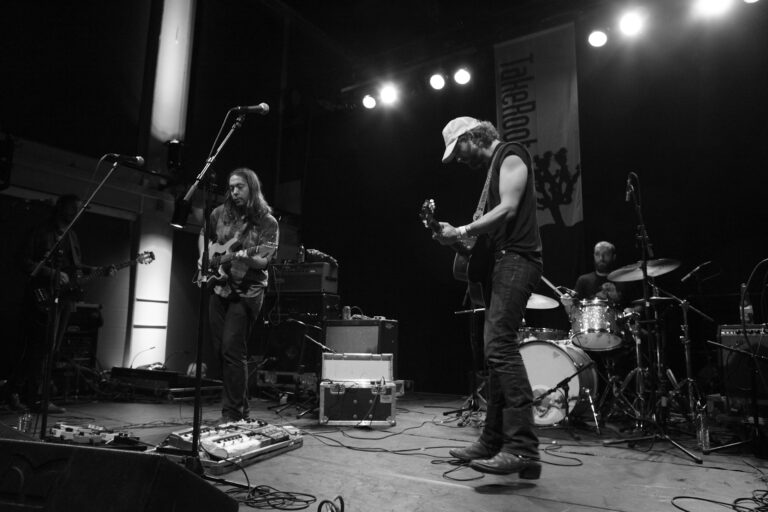Een band speelt live op een podium met een gitarist en een bassist, terwijl een drummer op de achtergrond zichtbaar is.