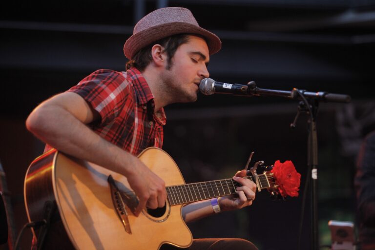 Een man met een hoed speelt gitaar en zingt in een microfoon op een podium.