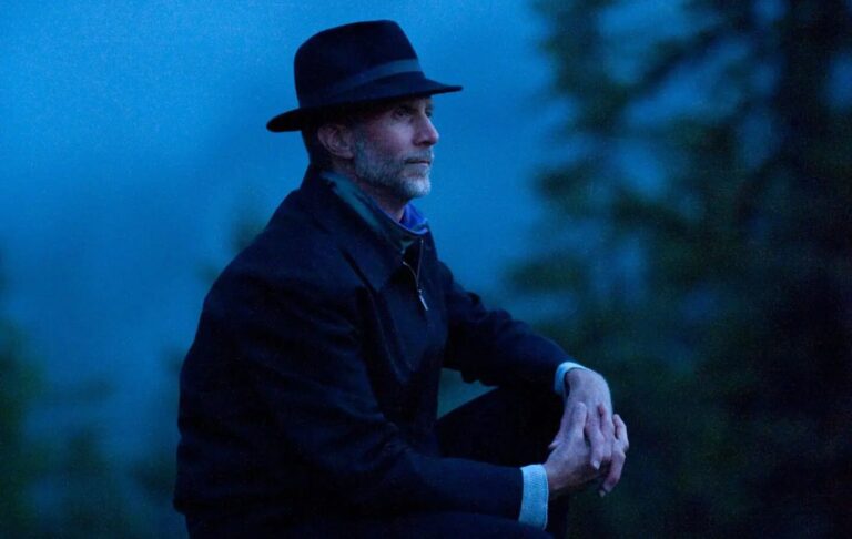 Een man in een zwart hoed en een donkere jas zit op zijn hurken in een blauwe, mistige omgeving.