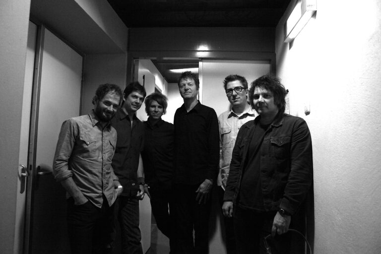 Een groep van zes mannen poseert in een smalle gang, met een zwarte en witte kleurenschema.