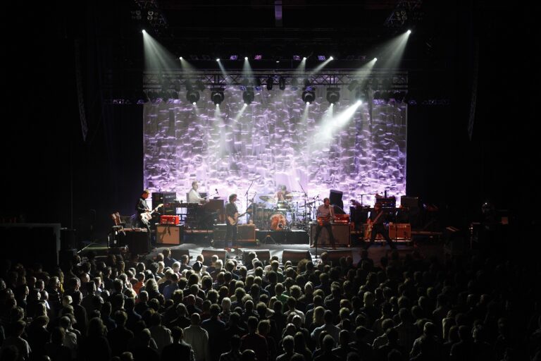 Een druk concert met een band op het podium en een groot publiek dat naar de show kijkt.