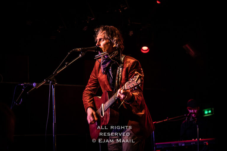 Een muzikant met een gitaar zingt op een podium, omringd door sfeervolle verlichting.