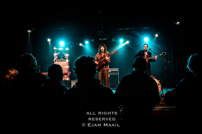 Een band speelt live op het podium met blauwe belichting, terwijl het publiek van achteren kijkt.