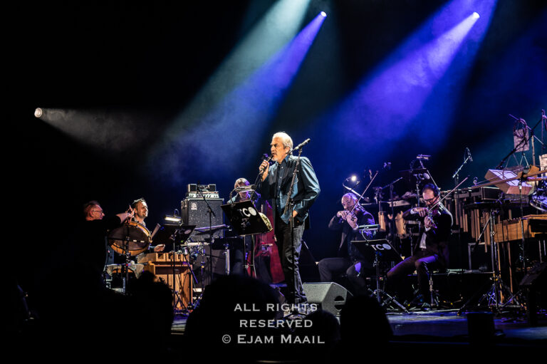 Een muzikant staat op het podium en zingt, omringd door een orkest met verschillende instrumenten en een kleurrijke lichtshow.
