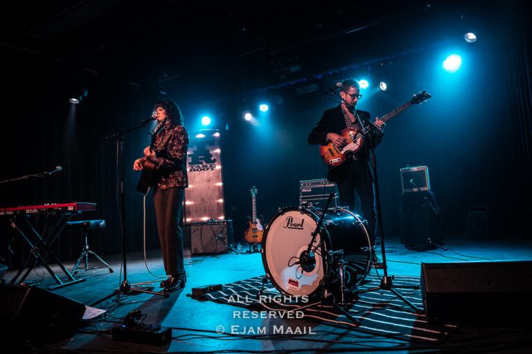 Twee muzikanten optreden op een podium met blauwe verlichting, met een gitaar en een drumstel zichtbaar.