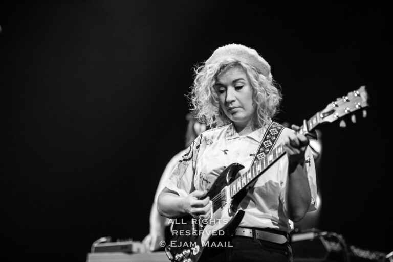Zangeres speelt gitaar op een podium met een donkere achtergrond.