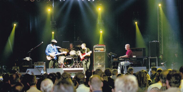 Een liveband speelt op een podium met felle lichten, terwijl het publiek naar hen kijkt.