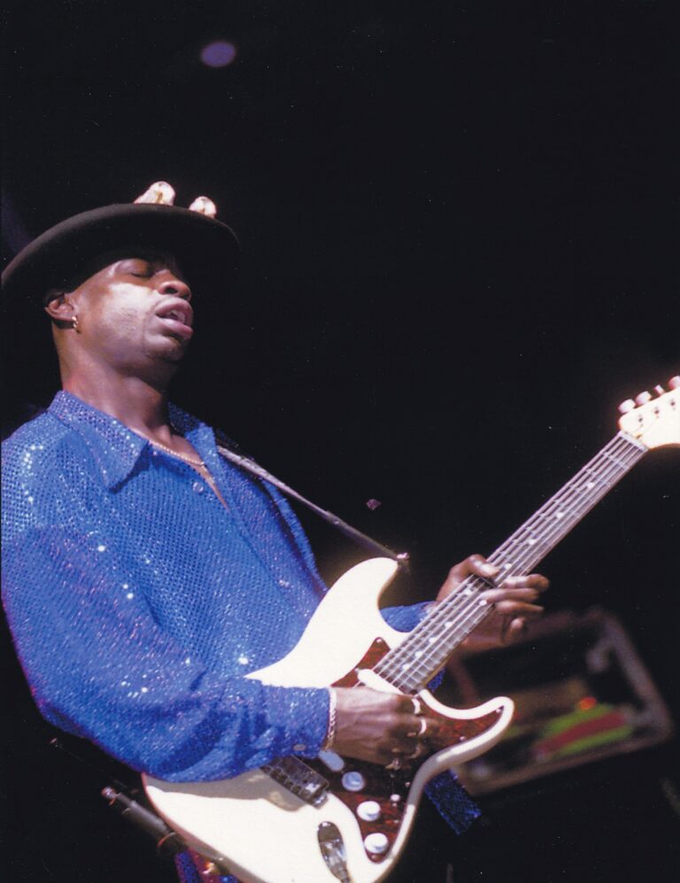 Een muzikant speelt gitaar op het podium, gekleed in een blauwe glinsterende blouse en een hoed.