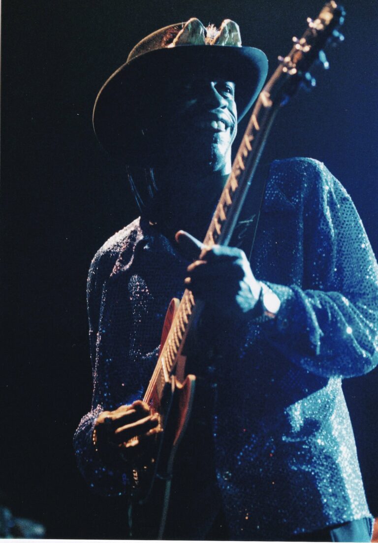 Een muzikant speelt gitaar op het podium, omgeven door een donkere achtergrond en lichtstralen.