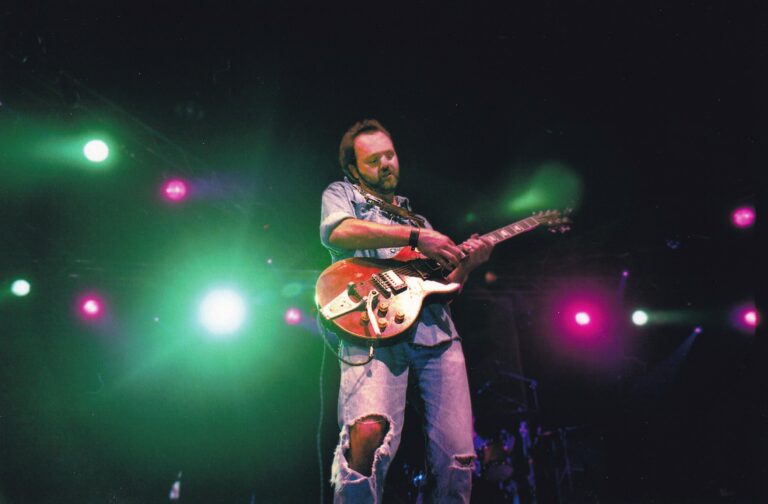 Een muzikant speelt gitaar op het podium met kleurrijke lichten op de achtergrond.