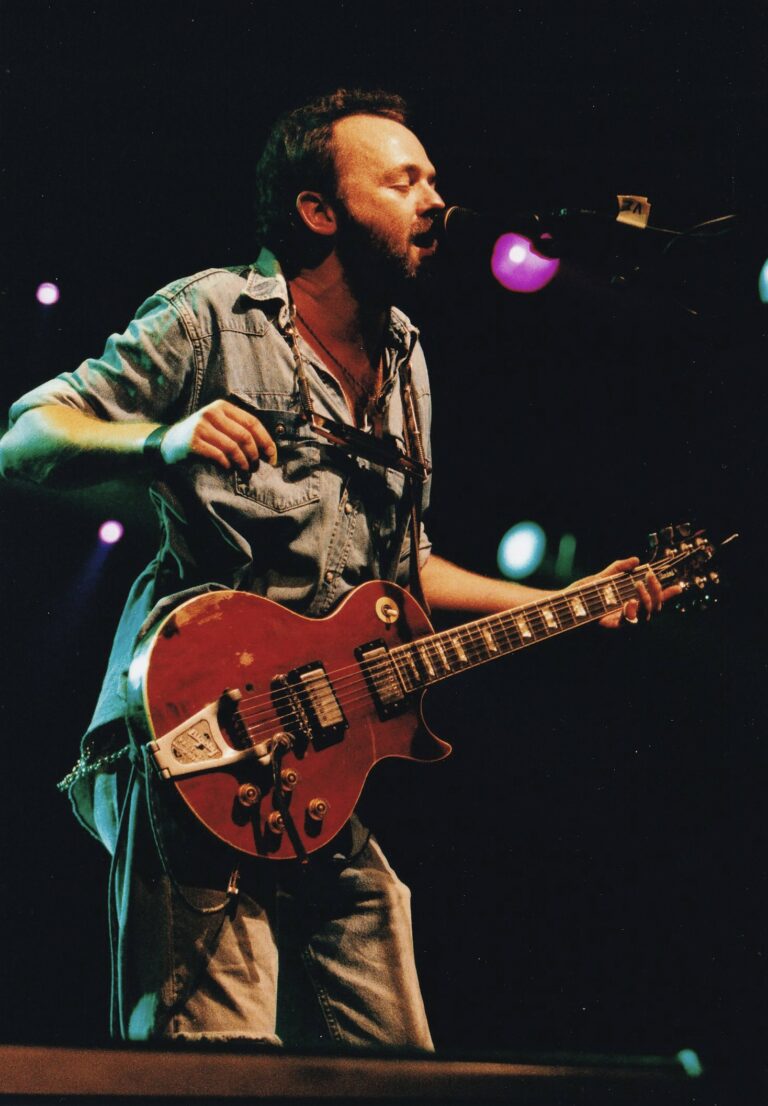 Een muzikant speelt gitaar op het podium met kleurrijke lichten op de achtergrond.