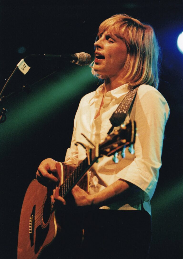 Een performer zingt in het licht van een podium met een gitaar in de hand.