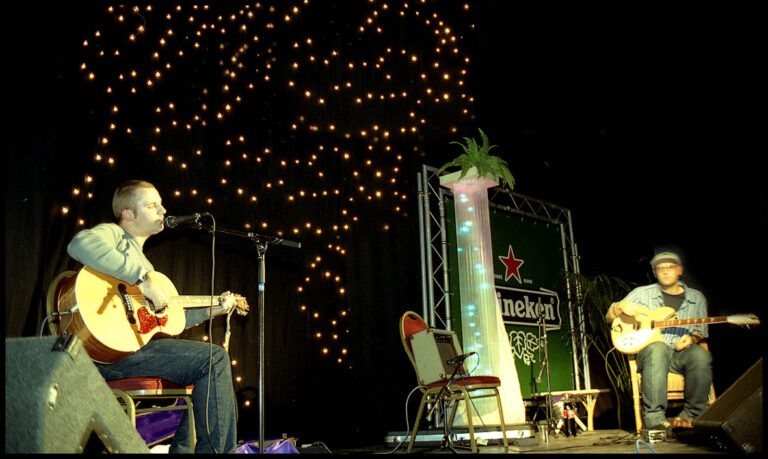 Een muzikant zit op een stoel en speelt gitaar op een podium versierd met sfeervolle verlichting.
