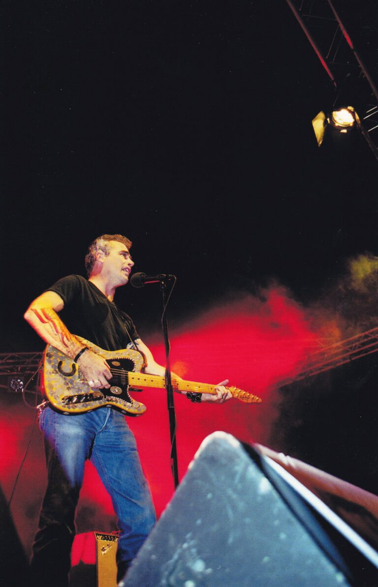 Een man staat op het podium met een gitaar, terwijl hij zingt onder een rode en zwarte achtergrond.