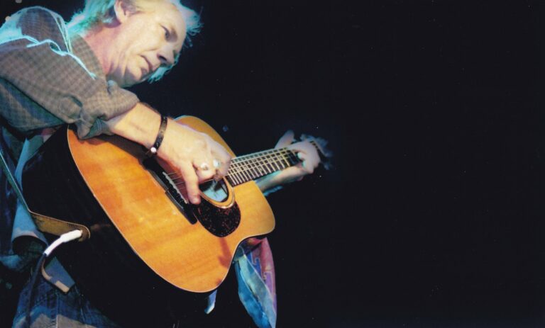 Een muzikant speelt een akoestische gitaar terwijl hij zich richt op zijn instrument in een donkere omgeving.