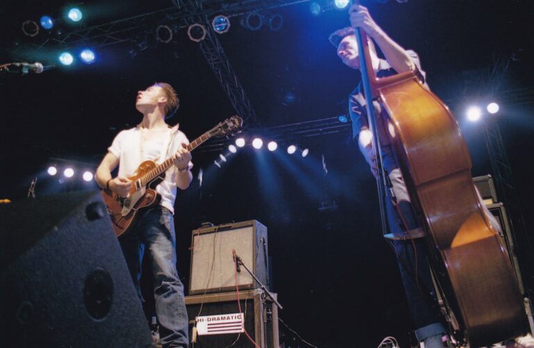 Twee muzikanten spelen live op een podium, met een elektrische gitaar en een contrabas, belicht door spotlights.
