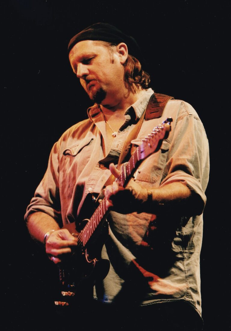 Man met een gitaar, geconcentreerd terwijl hij speelt op een podium met een donkere achtergrond.