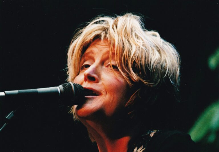 Een vrouw met kort, golvend haar zingt in een microfoon met een donkere achtergrond.