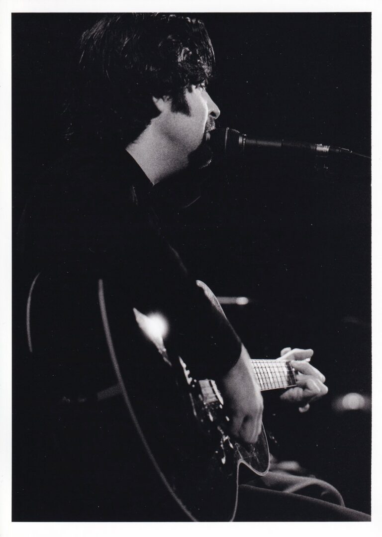 Zwart-wit afbeelding van een muzikant die een gitaar speelt en in een microfoon zingt.