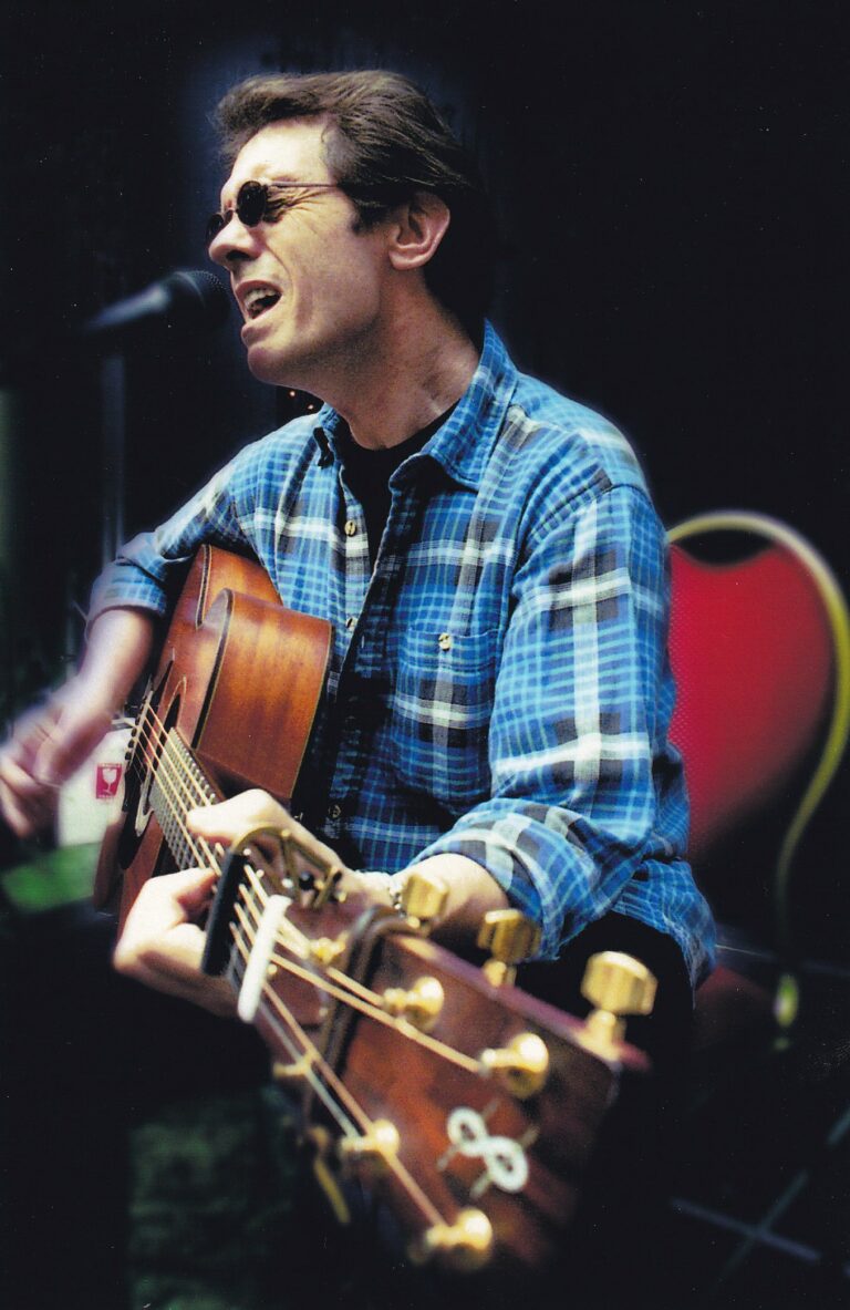 Een man in een blauw geruite overhemden die gitaar speelt en zingt op een podium.