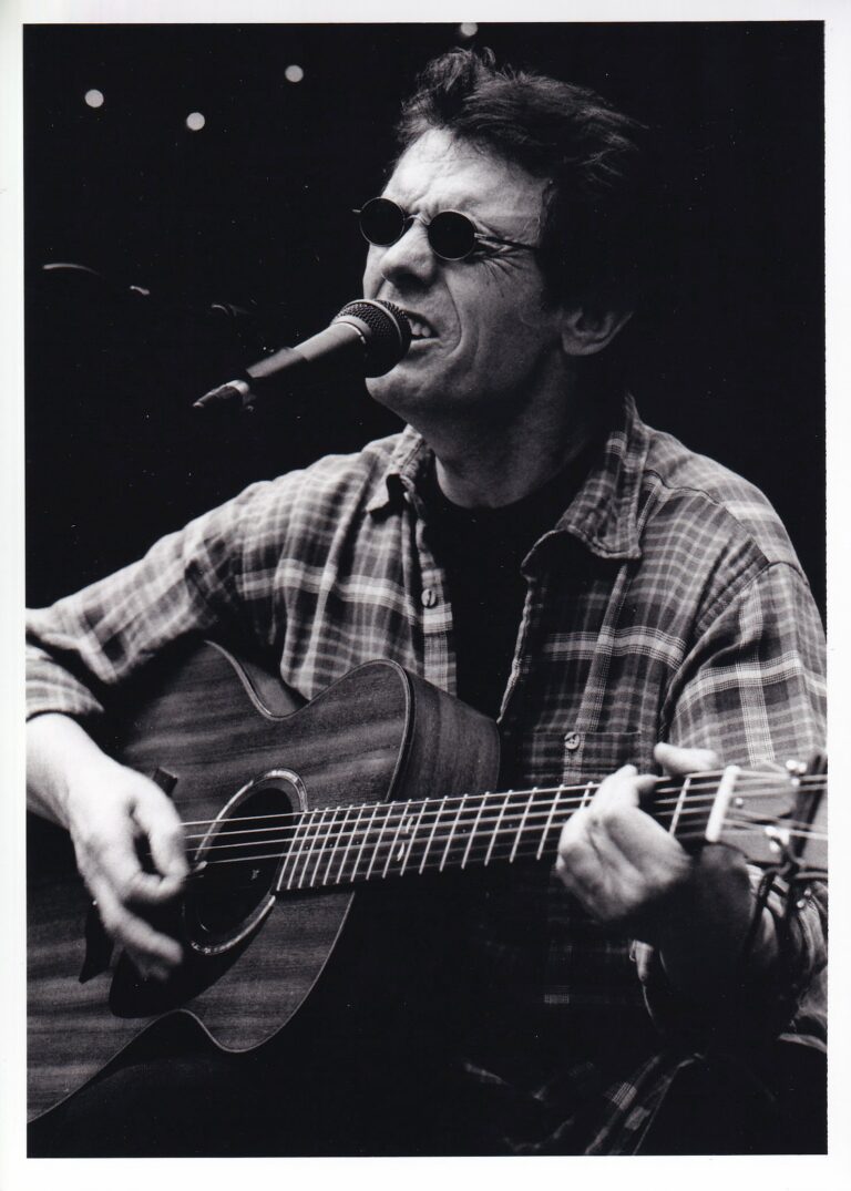 Een man met een gitaar zingt in een microfoon, gekleed in een geruite shirt en donkere zonnebril.