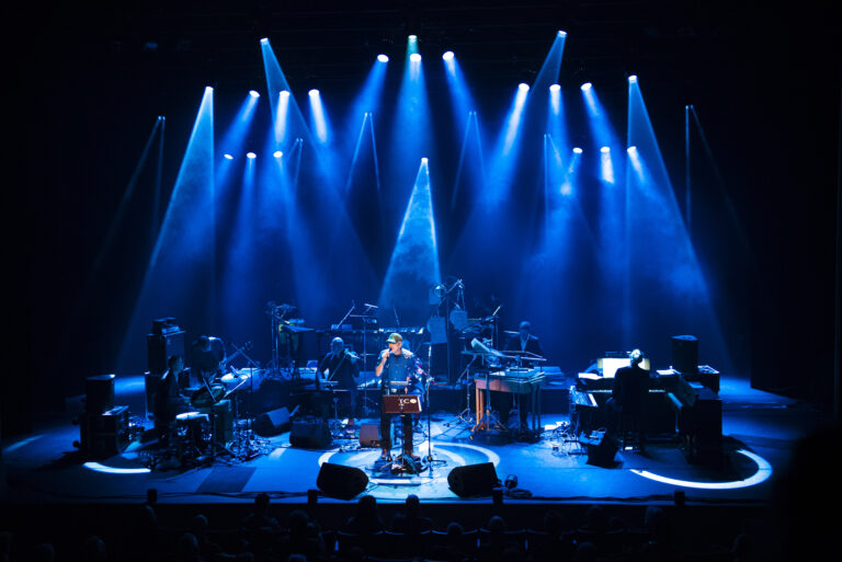 Een muzikant staat op het podium verlicht door blauwe lichten, omringd door verschillende instrumenten en muzikanten.