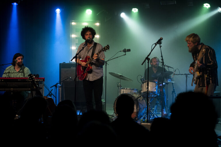 Een muzikant speelt gitaar op het podium terwijl een drummer op de achtergrond optreedt, verlicht door kleurrijke lichten.