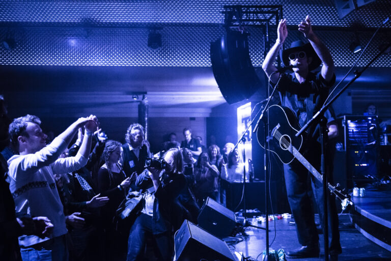 Een muzikant op het podium speelt gitaar terwijl het publiek enthousiast applaudisseert.