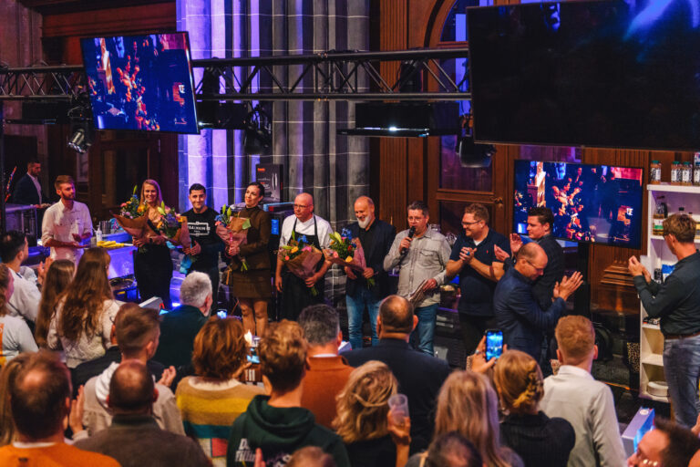 Een groep mensen staat op een podium in een feestelijke setting terwijl het publiek applaudisseert.