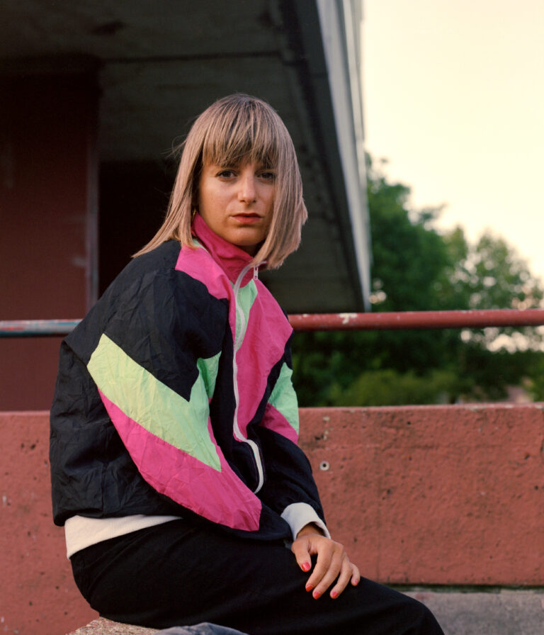 Een persoon met een korte, bleke haardos draagt een kleurrijke sportjack en zit op een bankje in een openluchtomgeving.